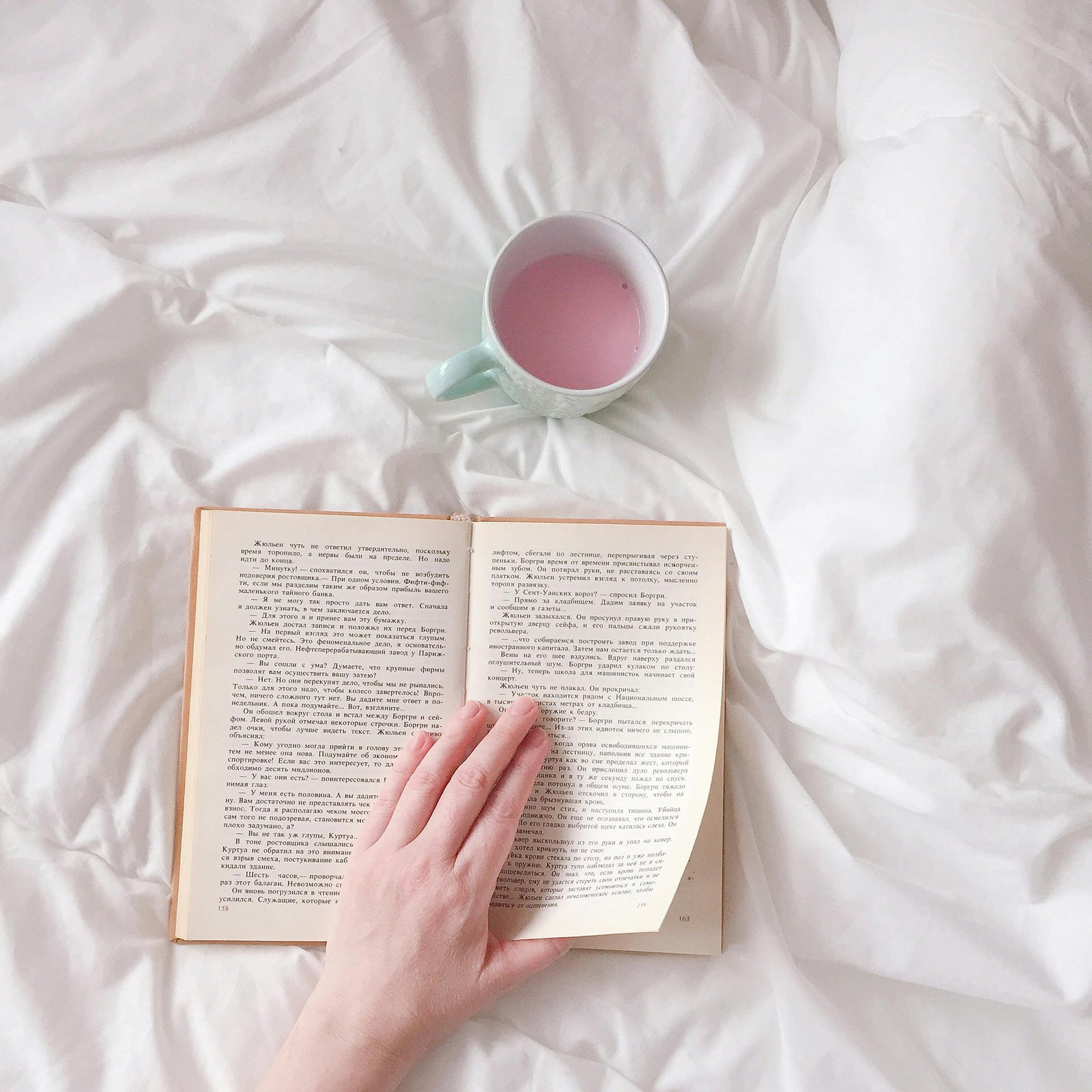 Relaxing scene of a hand turning book pages with a cup on white bedding, perfect for cozy reading vibes.